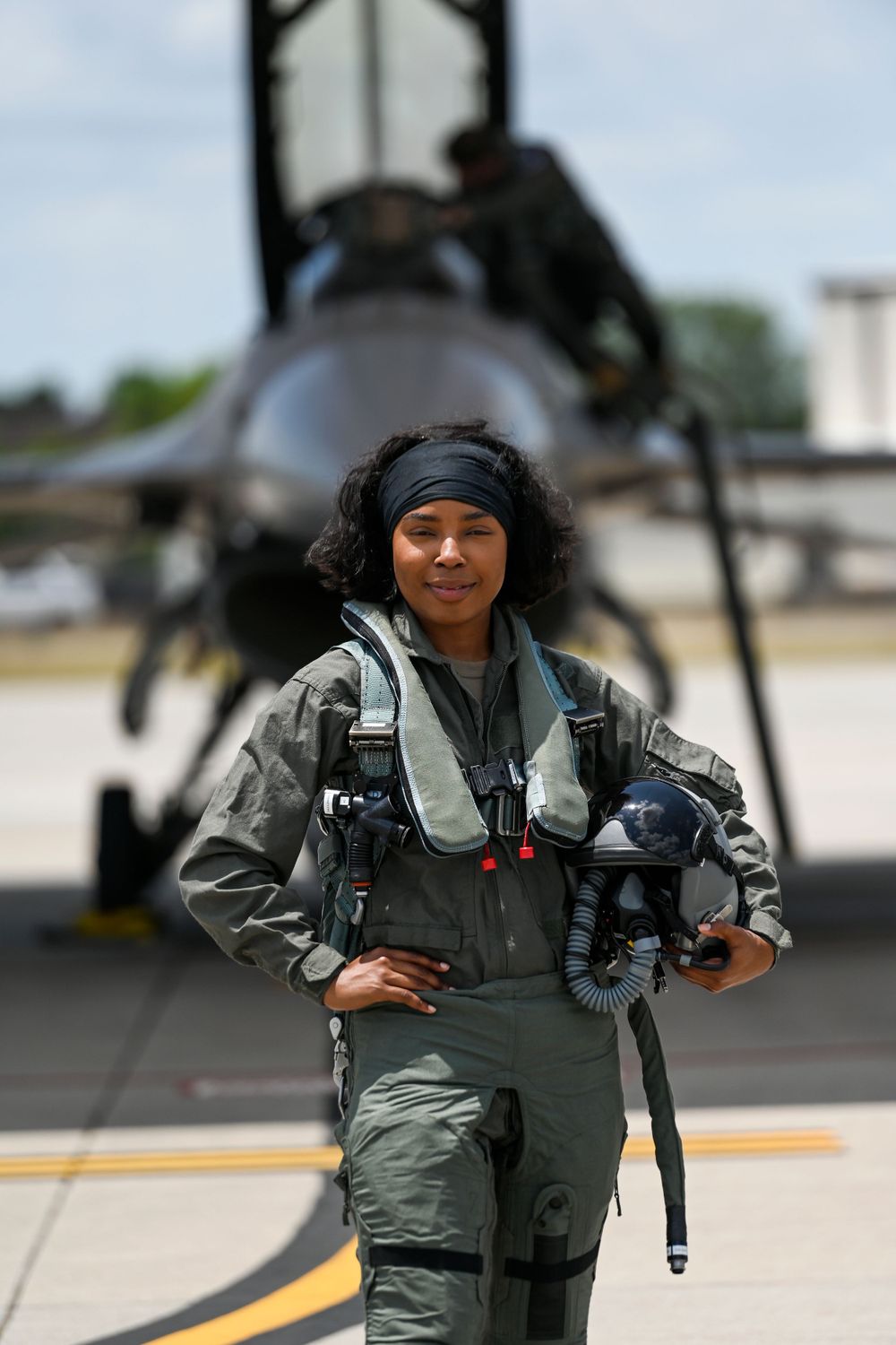 Military pilot in flight gear standing on airfield with aircraft in background.