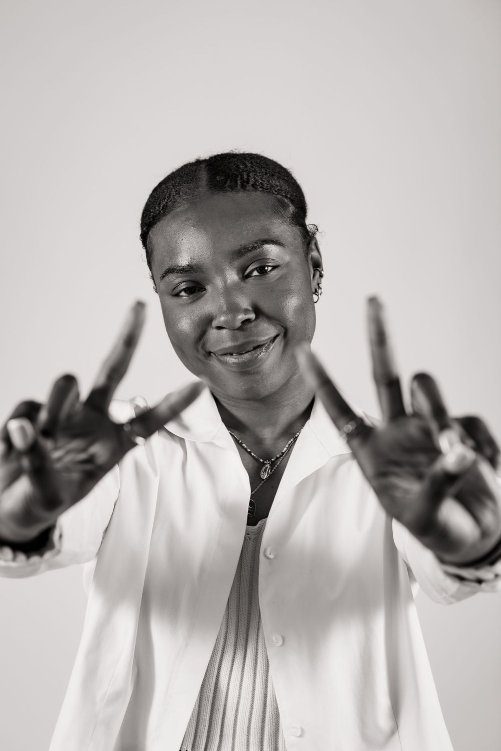 Someone wearing a white button-up shirt makes peace signs while smiling at the camera in black and white.