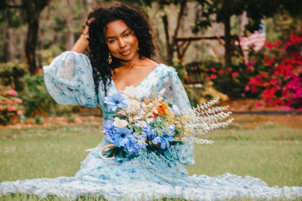 A person in a flowing light blue dress poses outdoors in a garden with vibrant flowers and lush greenery.
