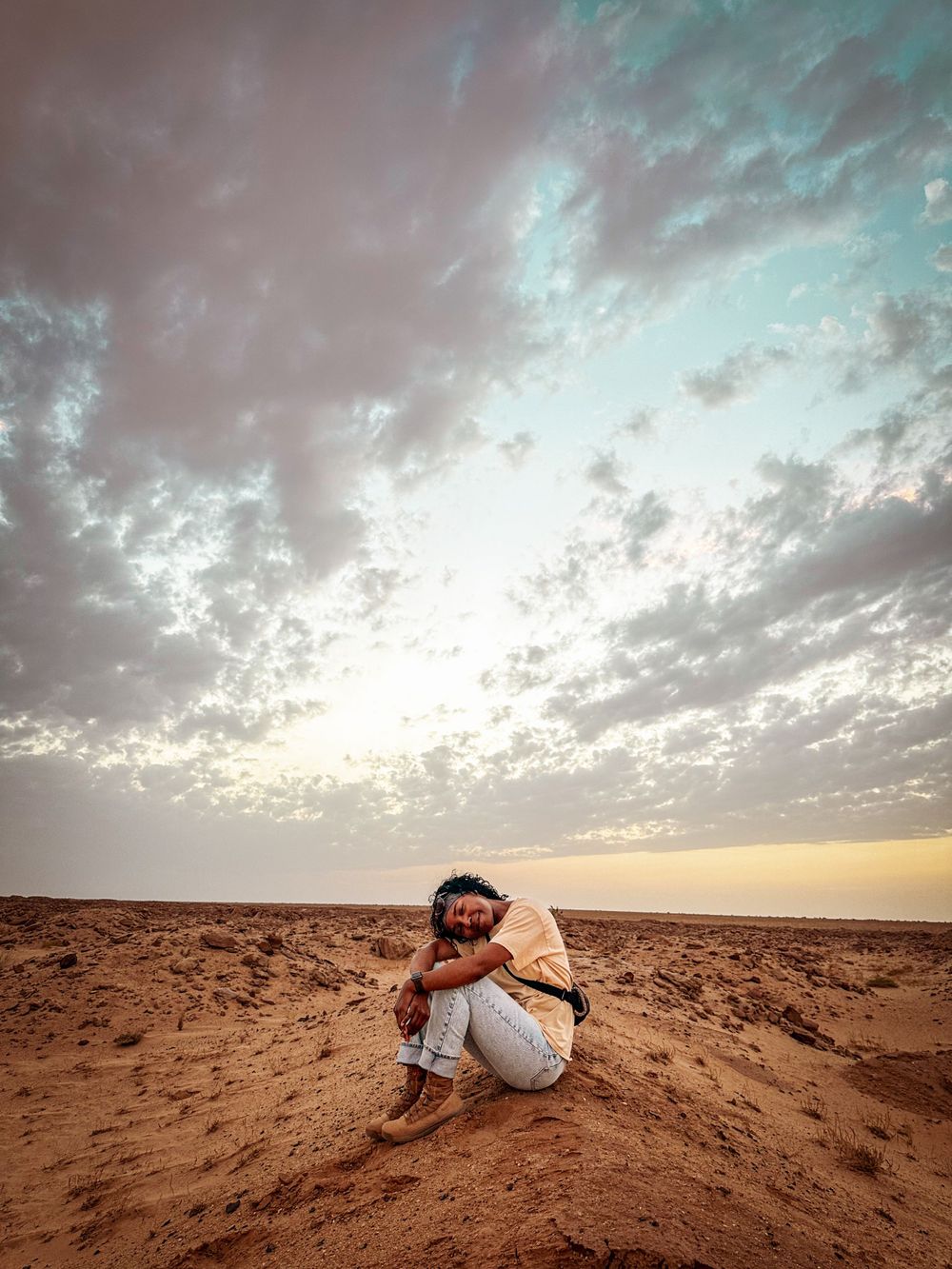 Person in traditional clothing crouching in desert landscape at sunset.