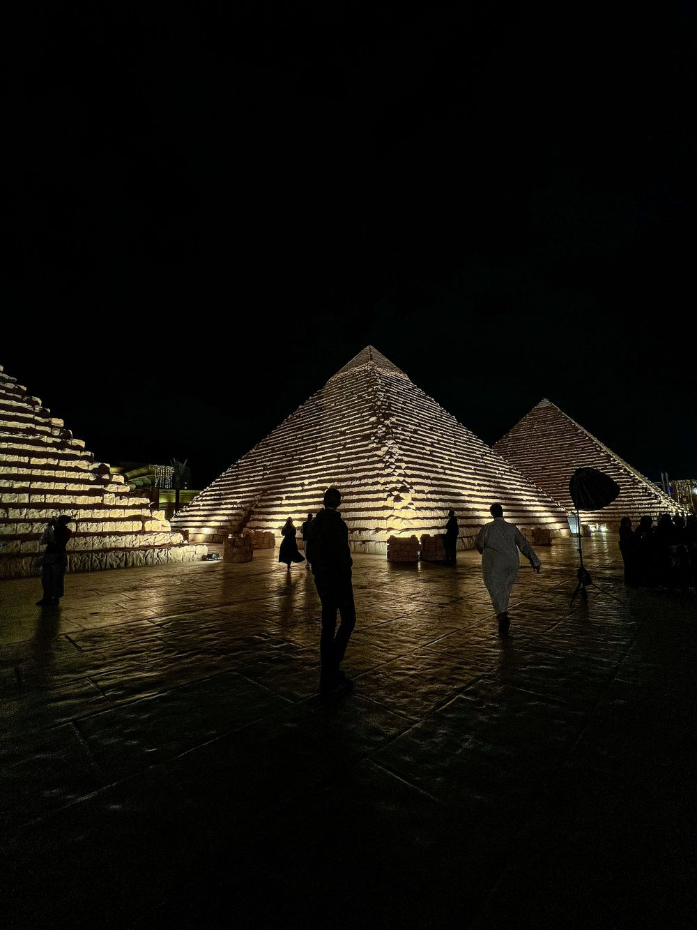 Illuminated pyramids of Egypt at night with silhouettes of visitors in the foreground.