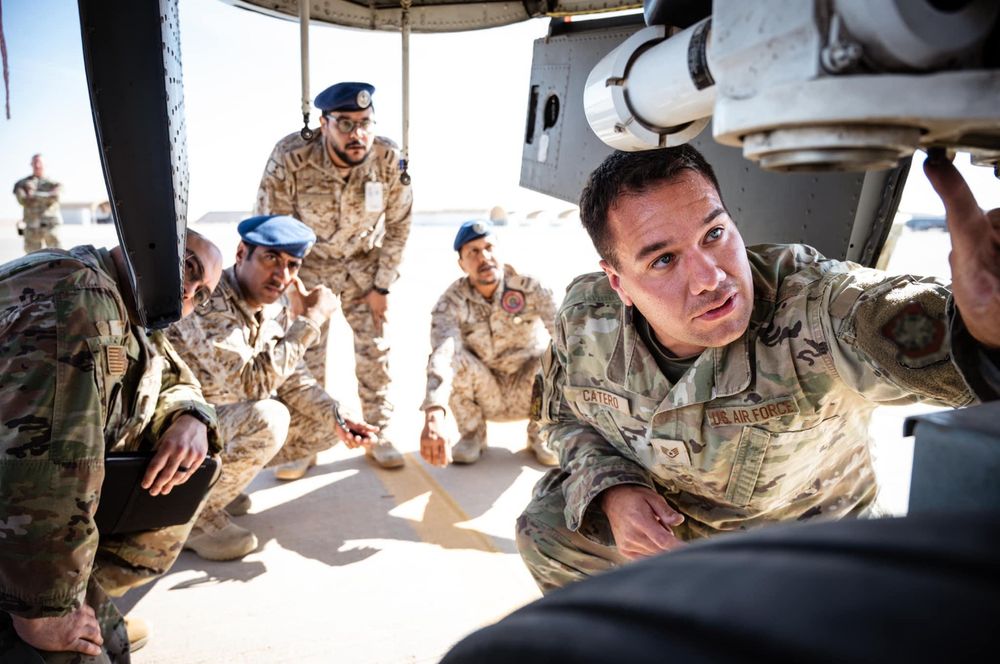 Group of military personnel gathered around a vehicle discussing mission details in desert conditions.