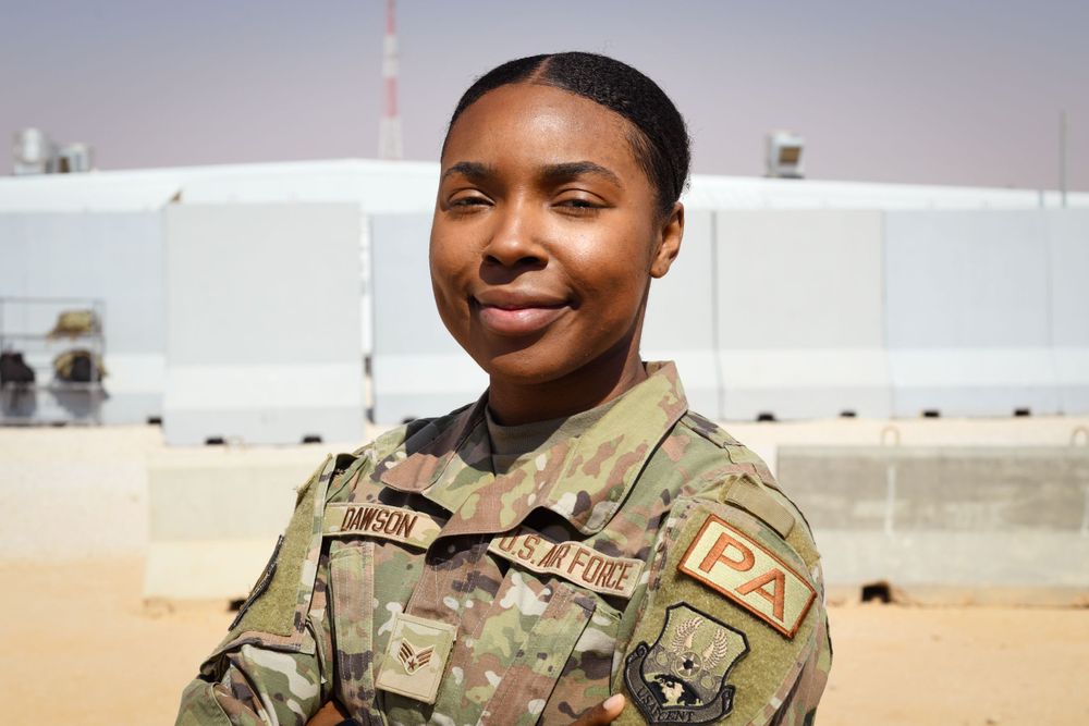 Service member in military camouflage uniform standing in front of white storage tanks.