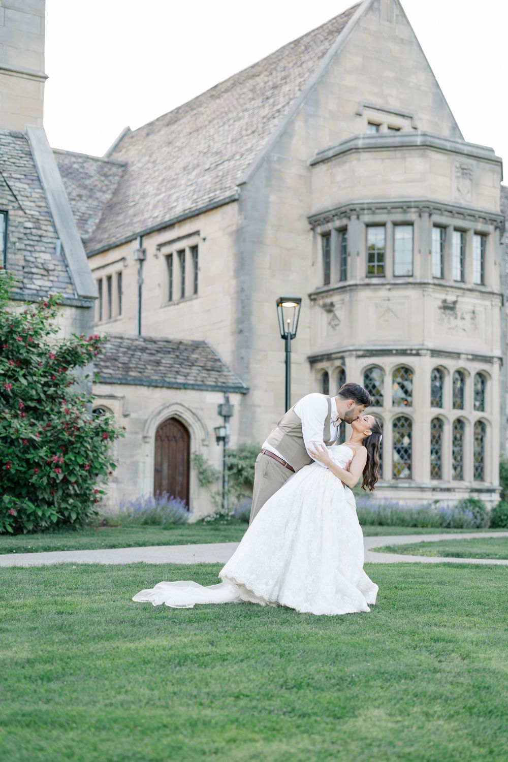 A bride and groom share a romantic dip kiss on a green lawn in front of an elegant stone manor house.