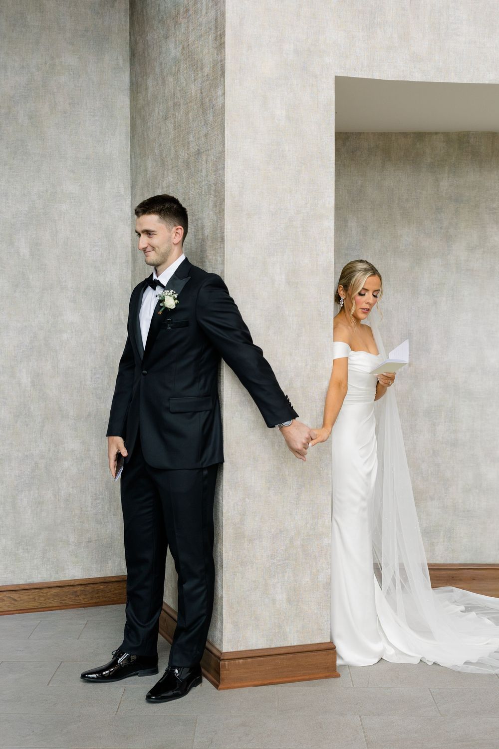 Bride in white off-shoulder gown reads vows while holding grooms hand around a pillar in elegant venue.