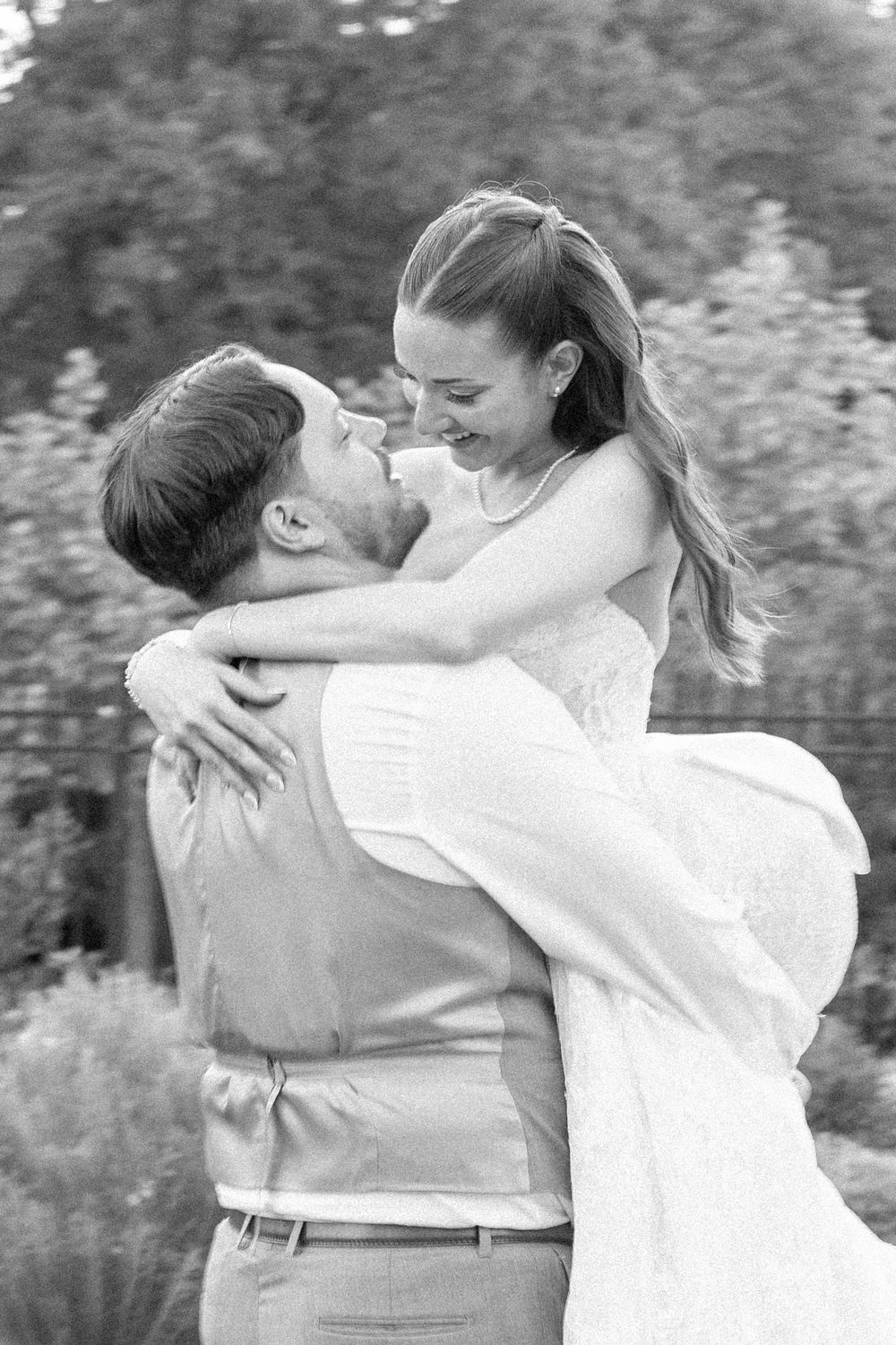 Black and white photo of a groom lifting his smiling bride in a wedding dress outdoors.