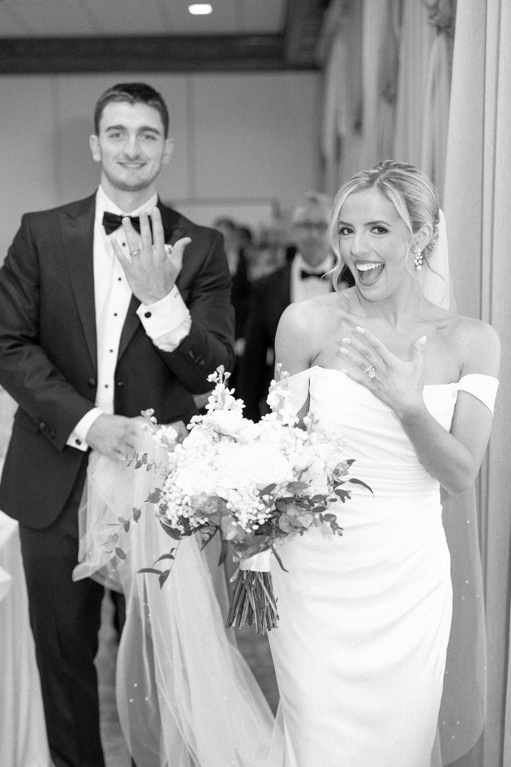Joyful bride and groom showing off wedding rings, bride holding bouquet in elegant white gown, black and white photo.