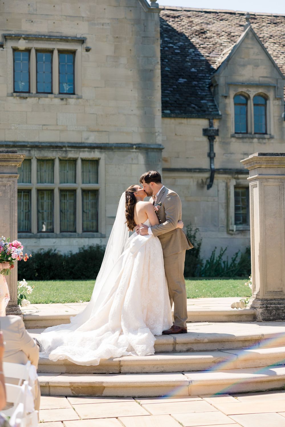 Bride and groom share first kiss on stone steps outside a grand English manor house on their wedding day.