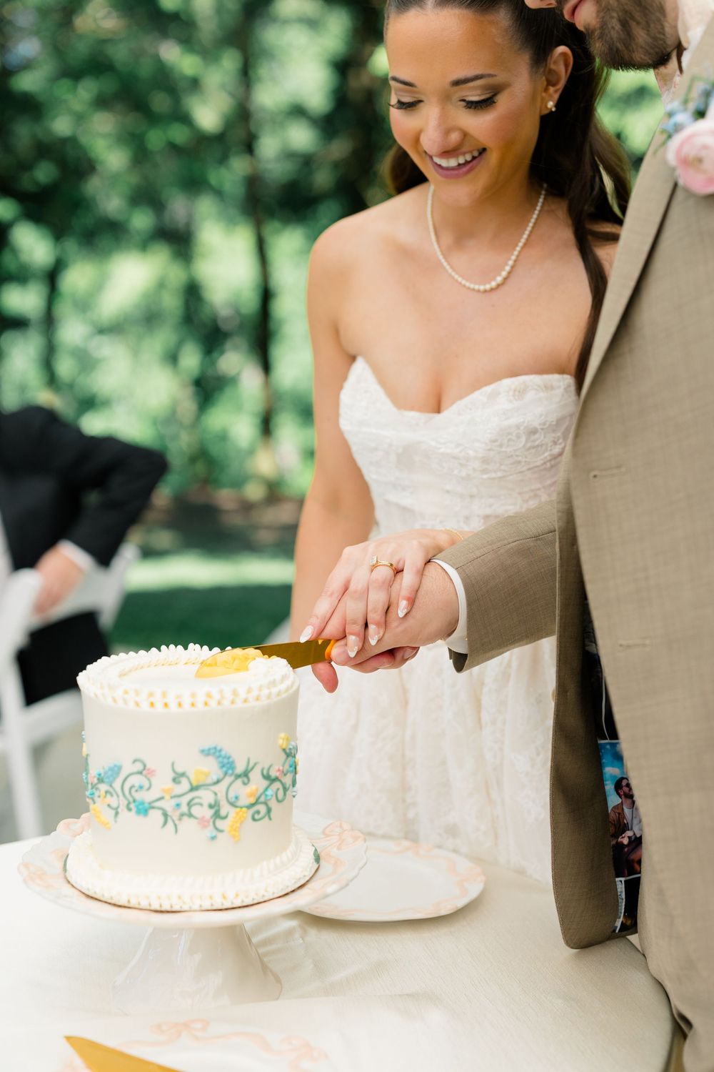 Smiling bride and groom cutting a white wedding cake with blue floral details outdoors.