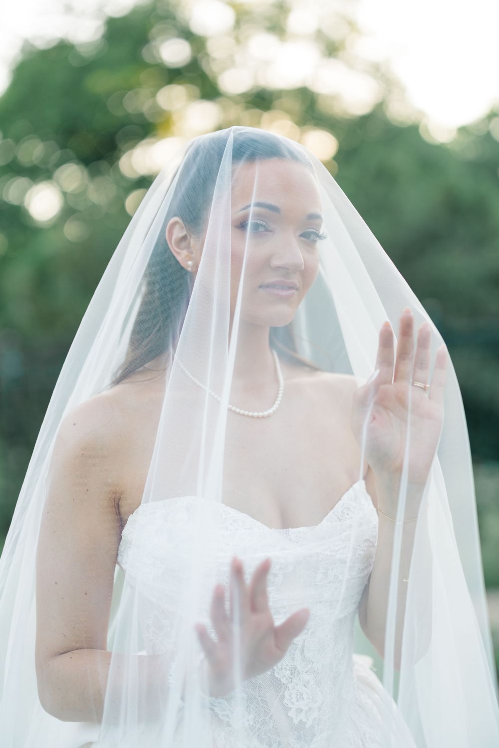 Bride in white lace gown and flowing veil holds fabric up, smiling softly outdoors with green bokeh background.