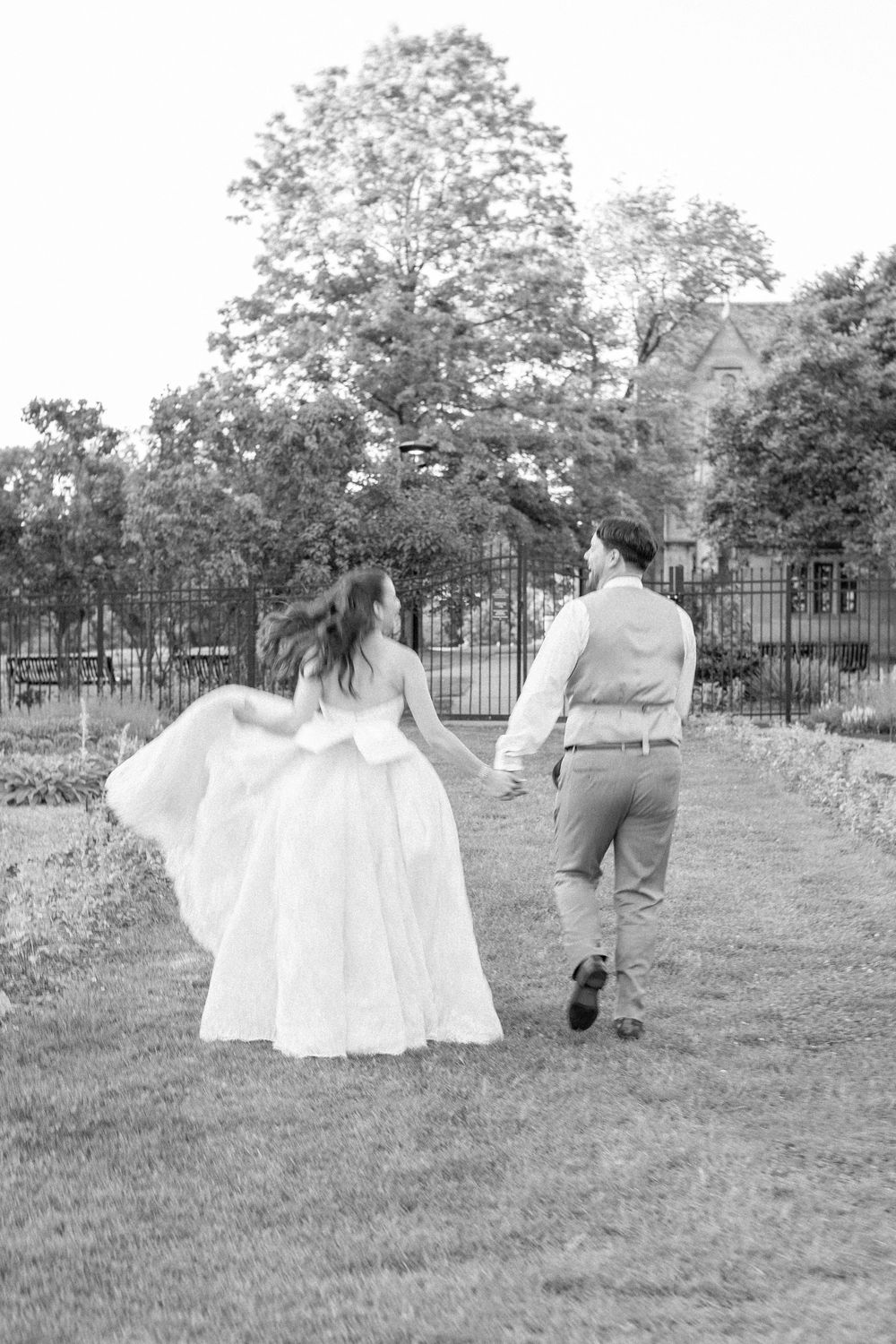 Black and white photo of bride and groom walking hand in hand through a garden path surrounded by trees.