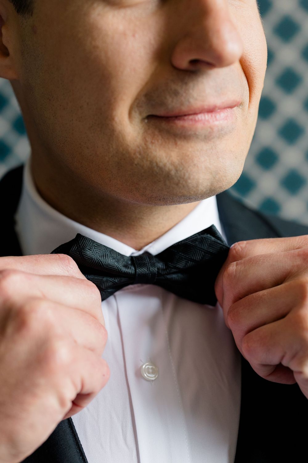 A groom in a black tuxedo adjusts his black bow tie, smiling slightly against a blue patterned background.
