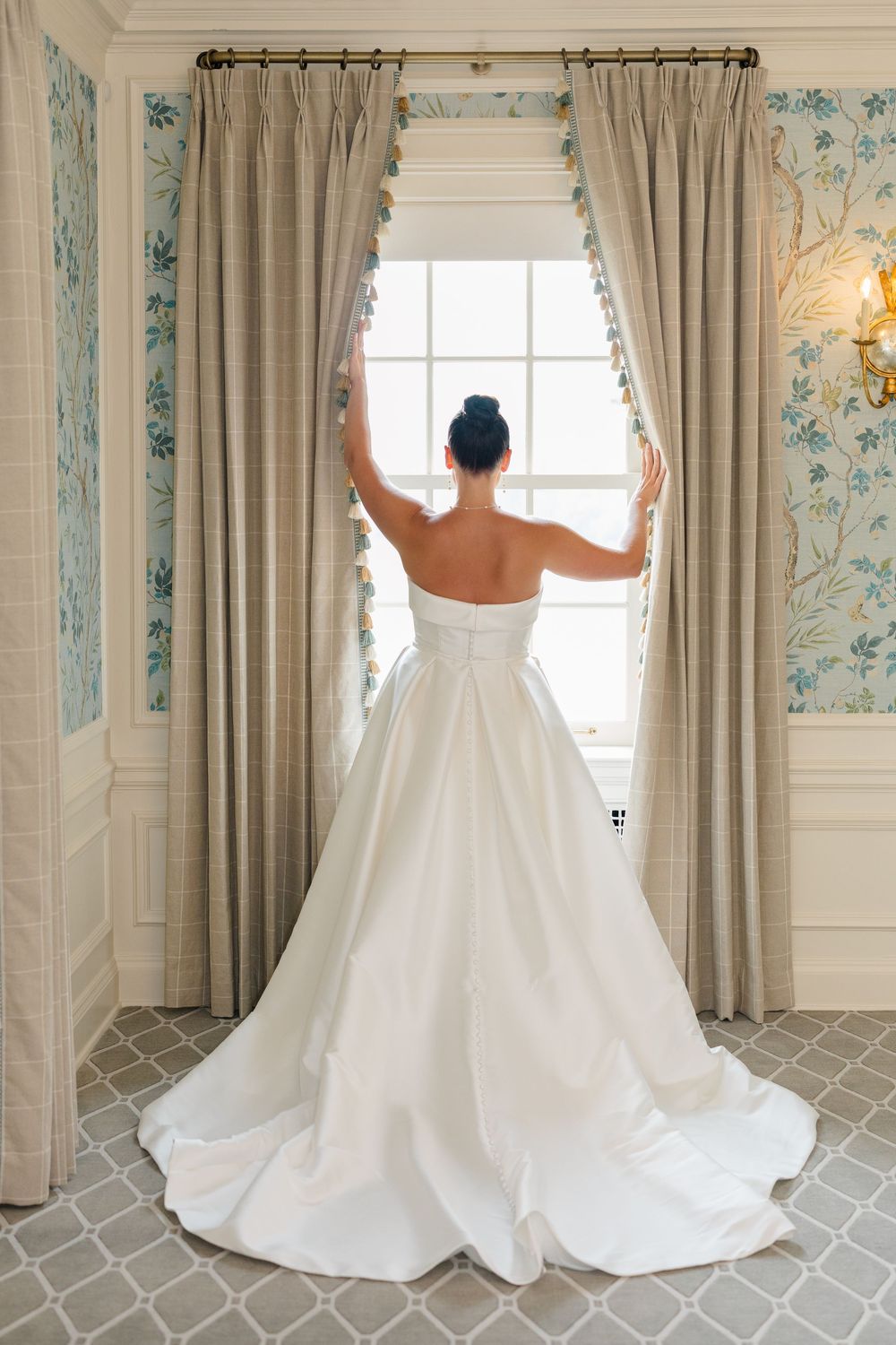 Bride in a strapless white ballgown with a long train stands facing a window, framed by elegant curtains and floral wallpaper.