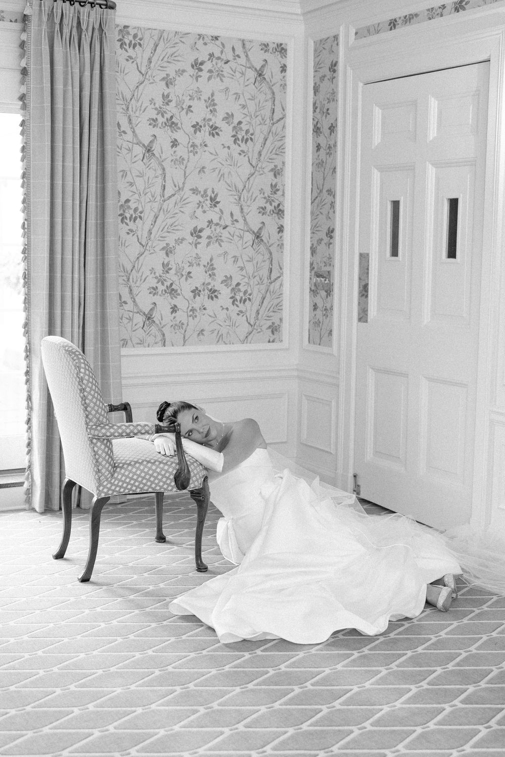 Bride in white gown sitting on floor leaning on chair in elegant room with floral wallpaper, black and white photo.