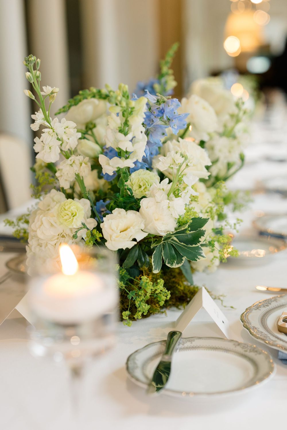 Elegant wedding reception table with white and blue floral centerpiece, candles, and fine china place settings.