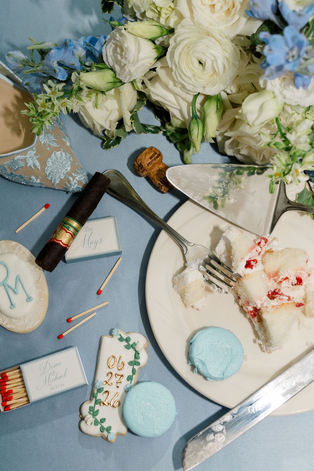 Elegant wedding flat lay with white flowers, cake slice, macarons, matchbooks, and a cigar on a blue surface.