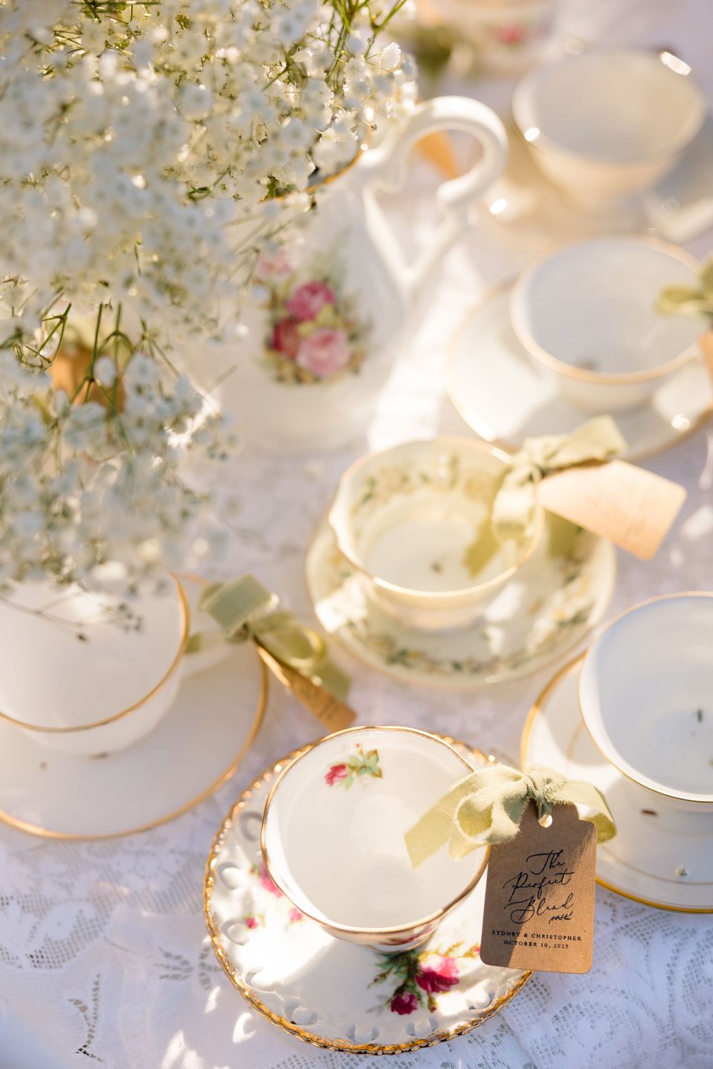 Elegant vintage floral teacups with gold rims and ribbon tags arranged on lace tablecloth with baby's breath.