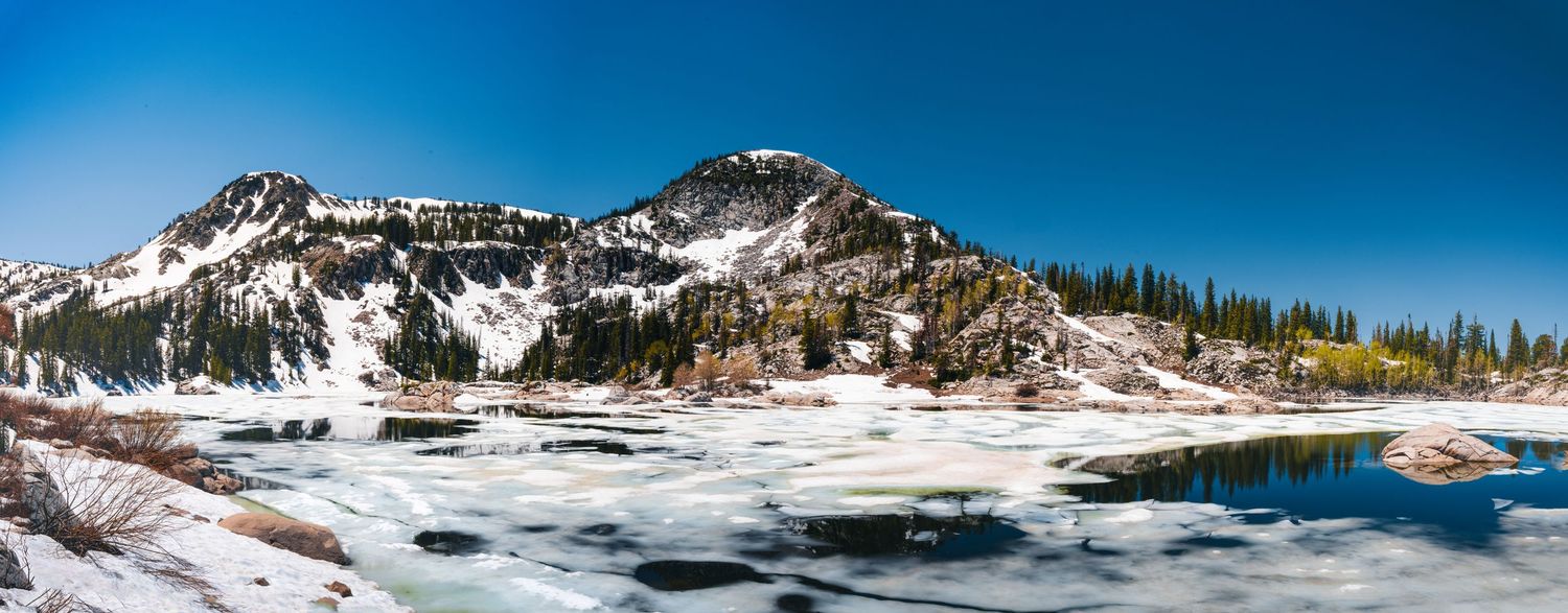Snow-capped mountain peak reflects in partially frozen alpine lake under blue sky.