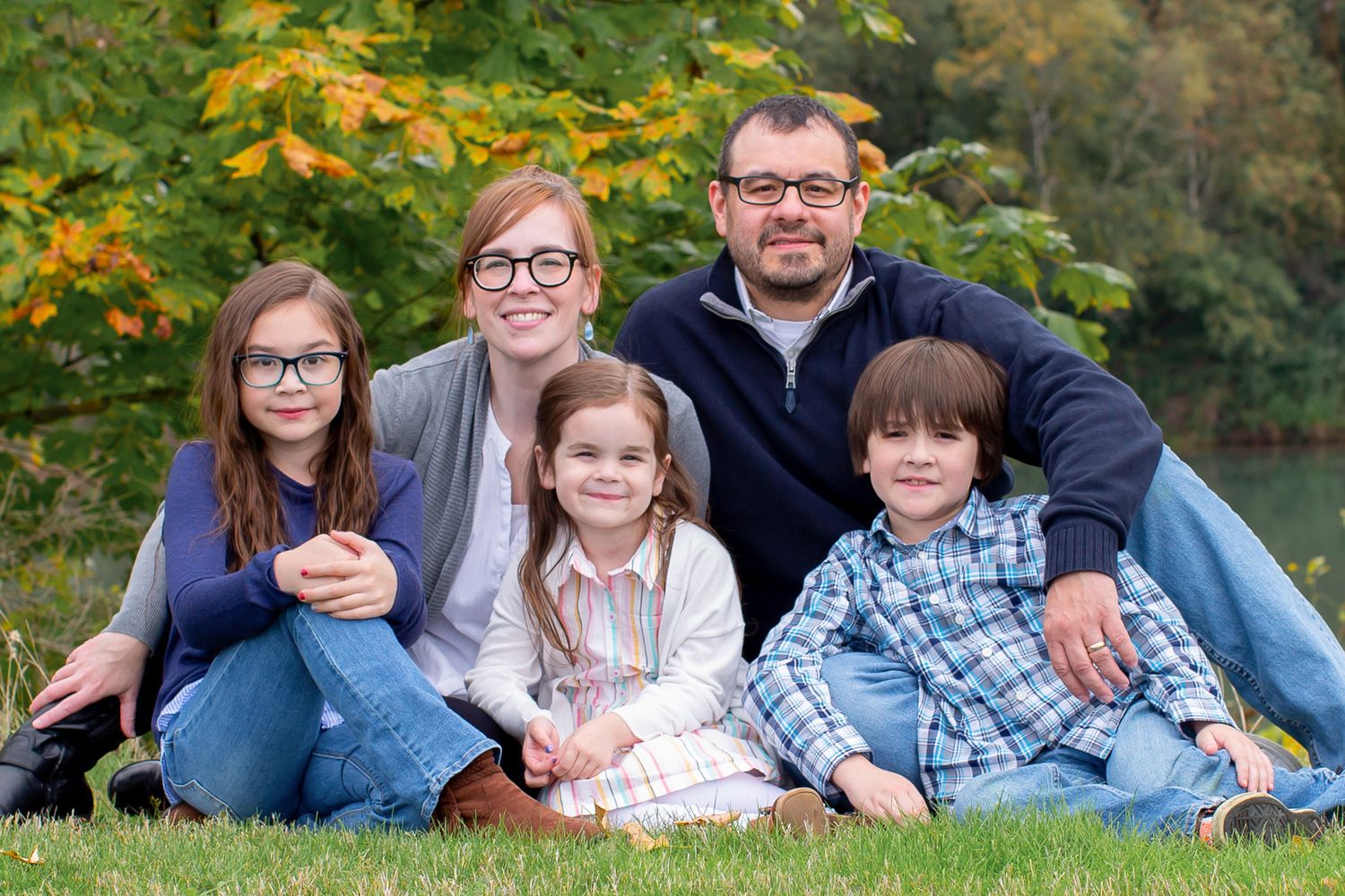 Family of five sitting close together on grass with autumn foliage background.