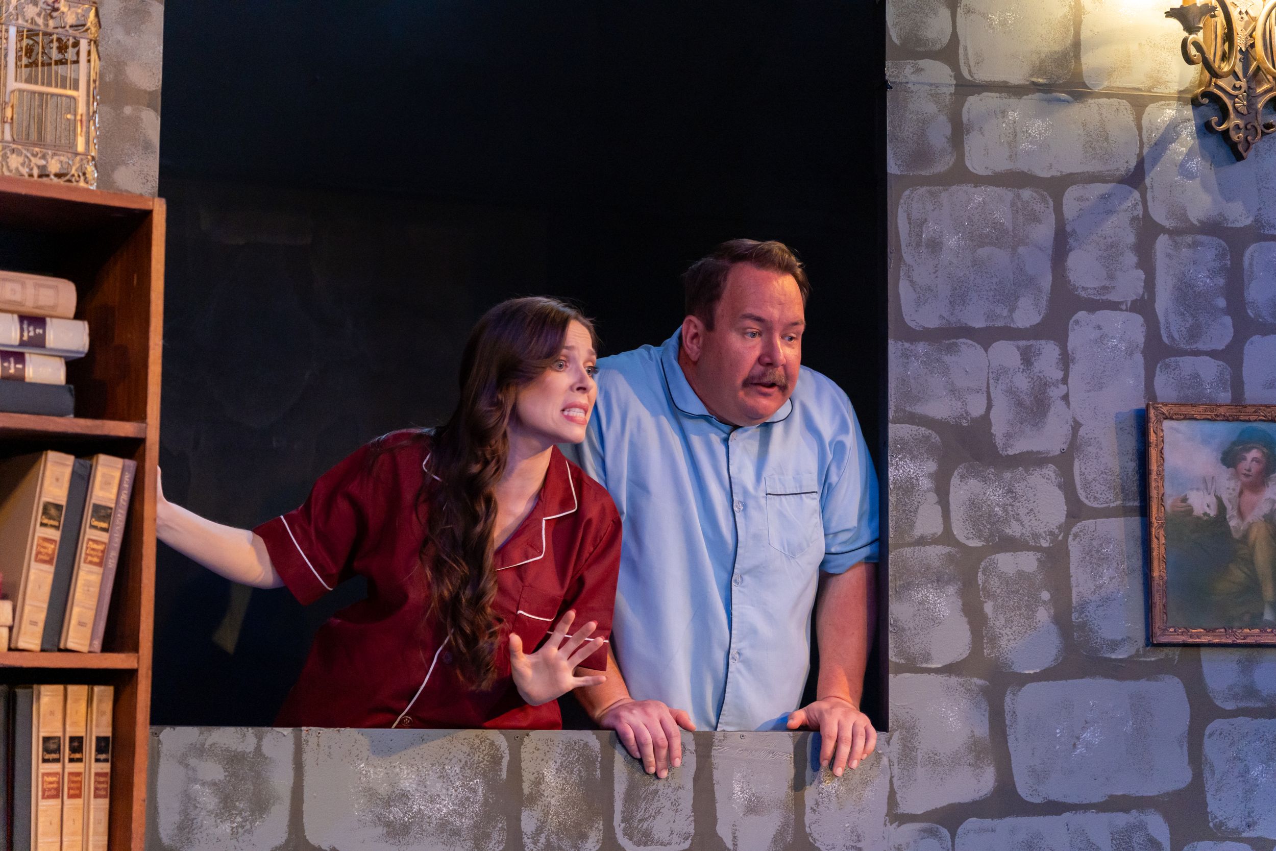 Two performers lean out from a stone window set piece during theatrical performance.