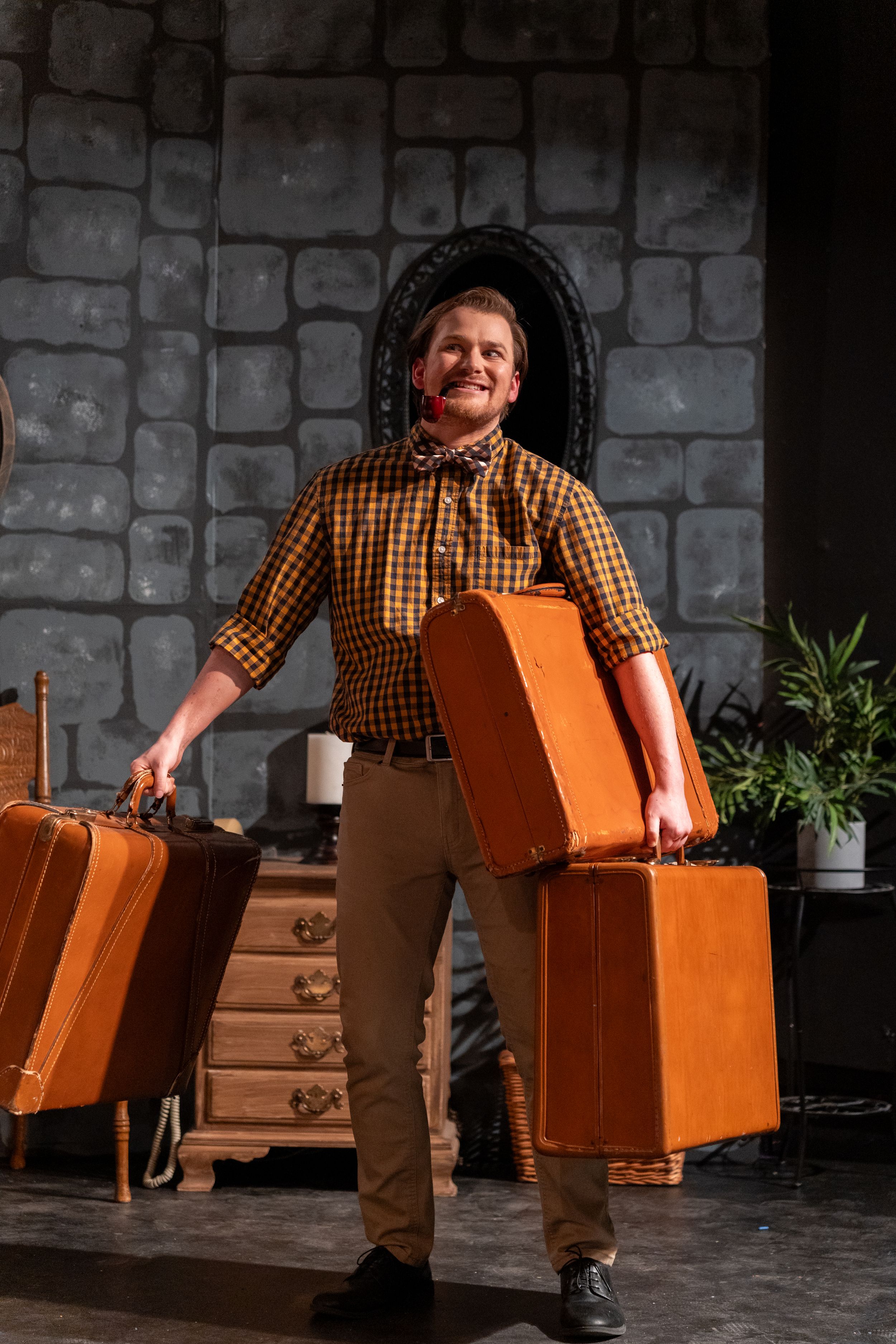 A performer carries orange suitcases while standing against a stone wall backdrop.