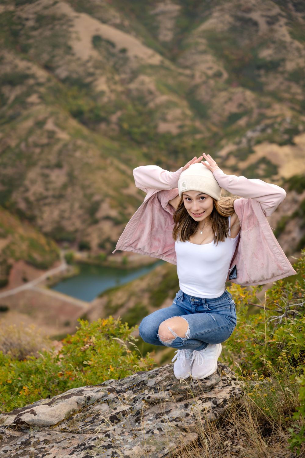 Adventurous mountain pose with scenic lake view wearing pink jacket and ripped jeans.