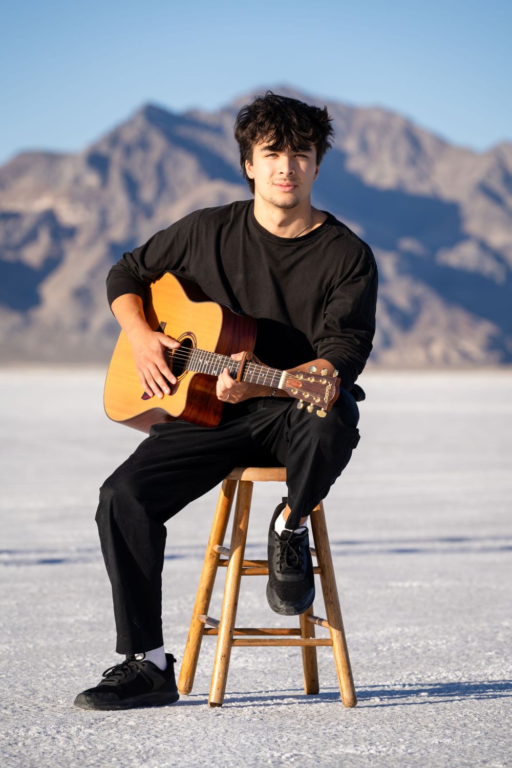 Guitarist in black outfit performs on wooden stool against mountain backdrop.