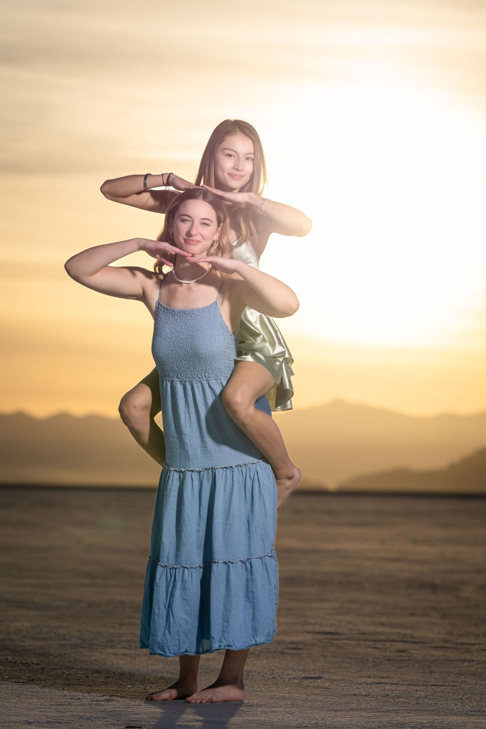 Two figures in flowing dresses pose playfully in desert landscape at sunset.