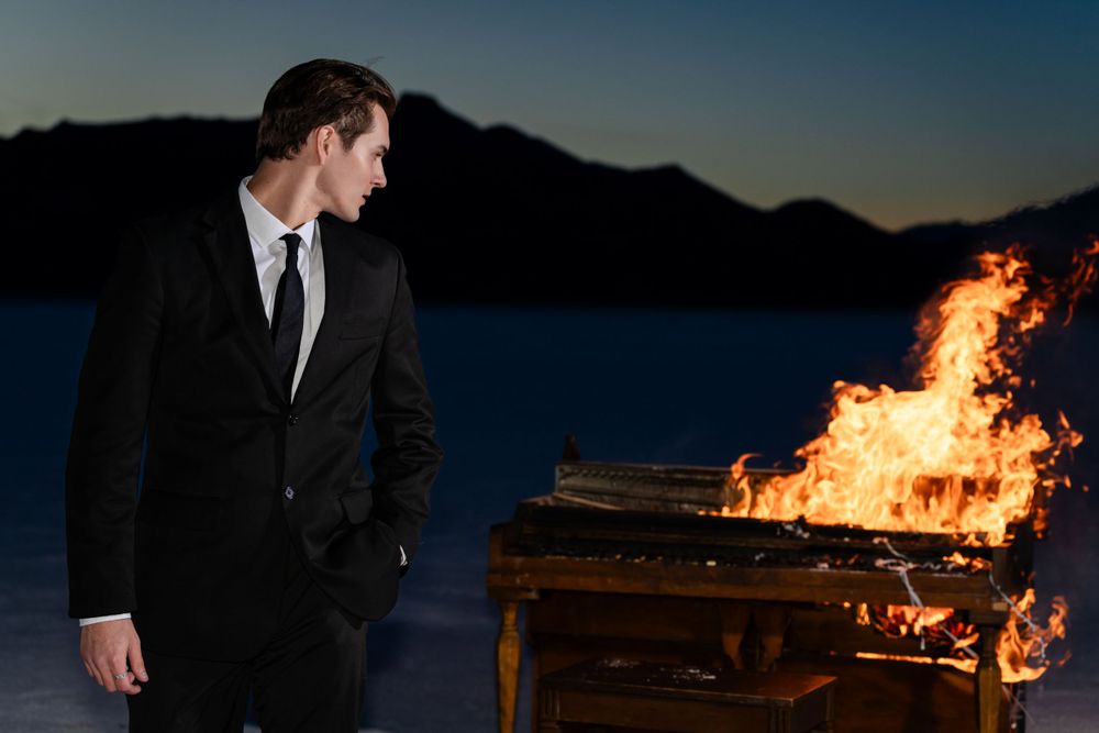 Formal suit against burning piano with mountains silhouetted at dusk.