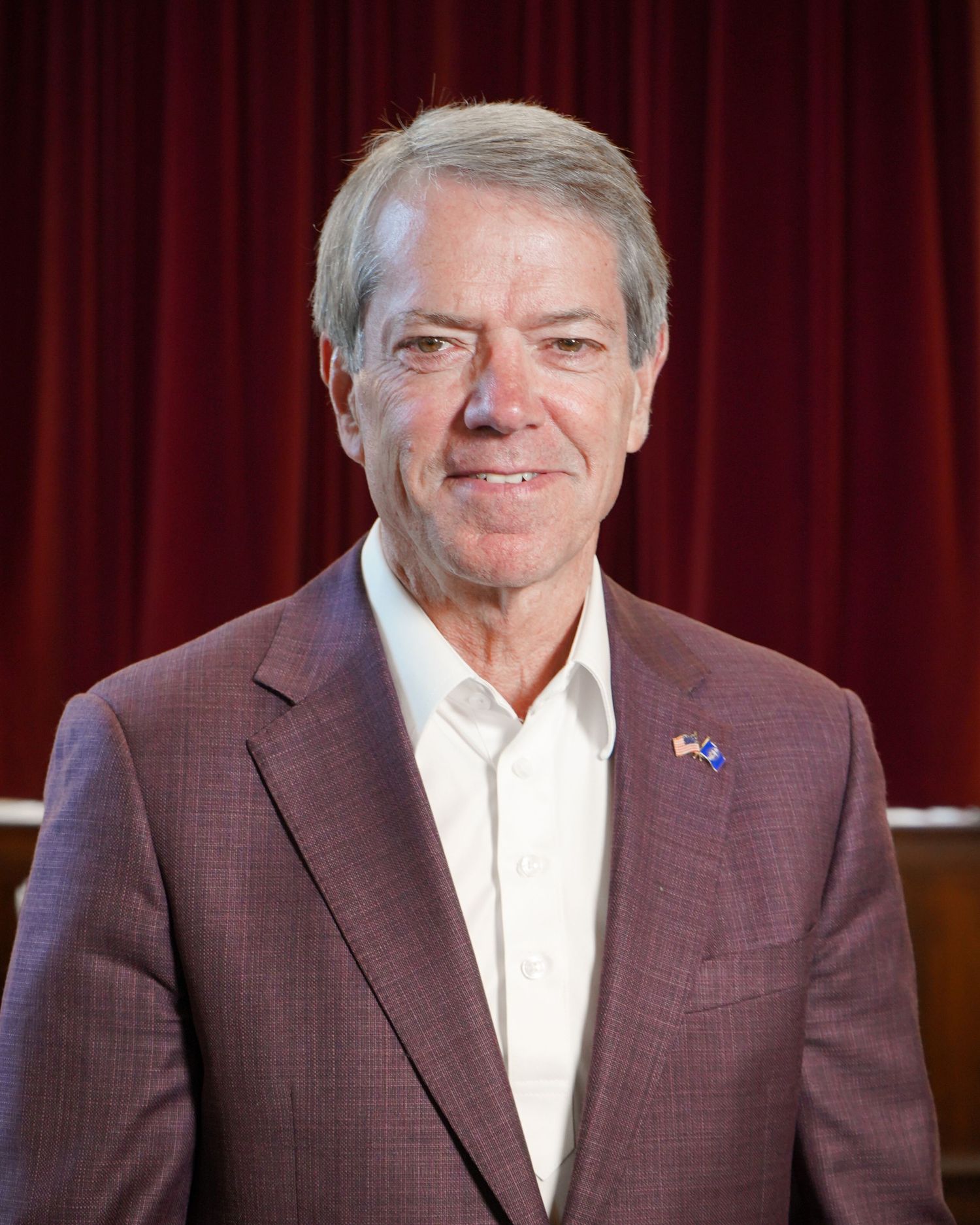 Professional portrait of senior executive in brown suit jacket against red curtain backdrop.