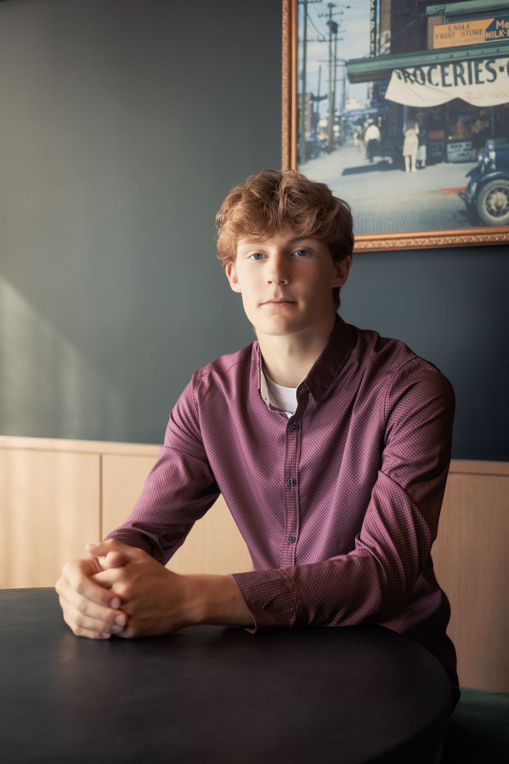 Young person seated at a table wearing a burgundy zip-up jacket in a dimly lit restaurant setting.
