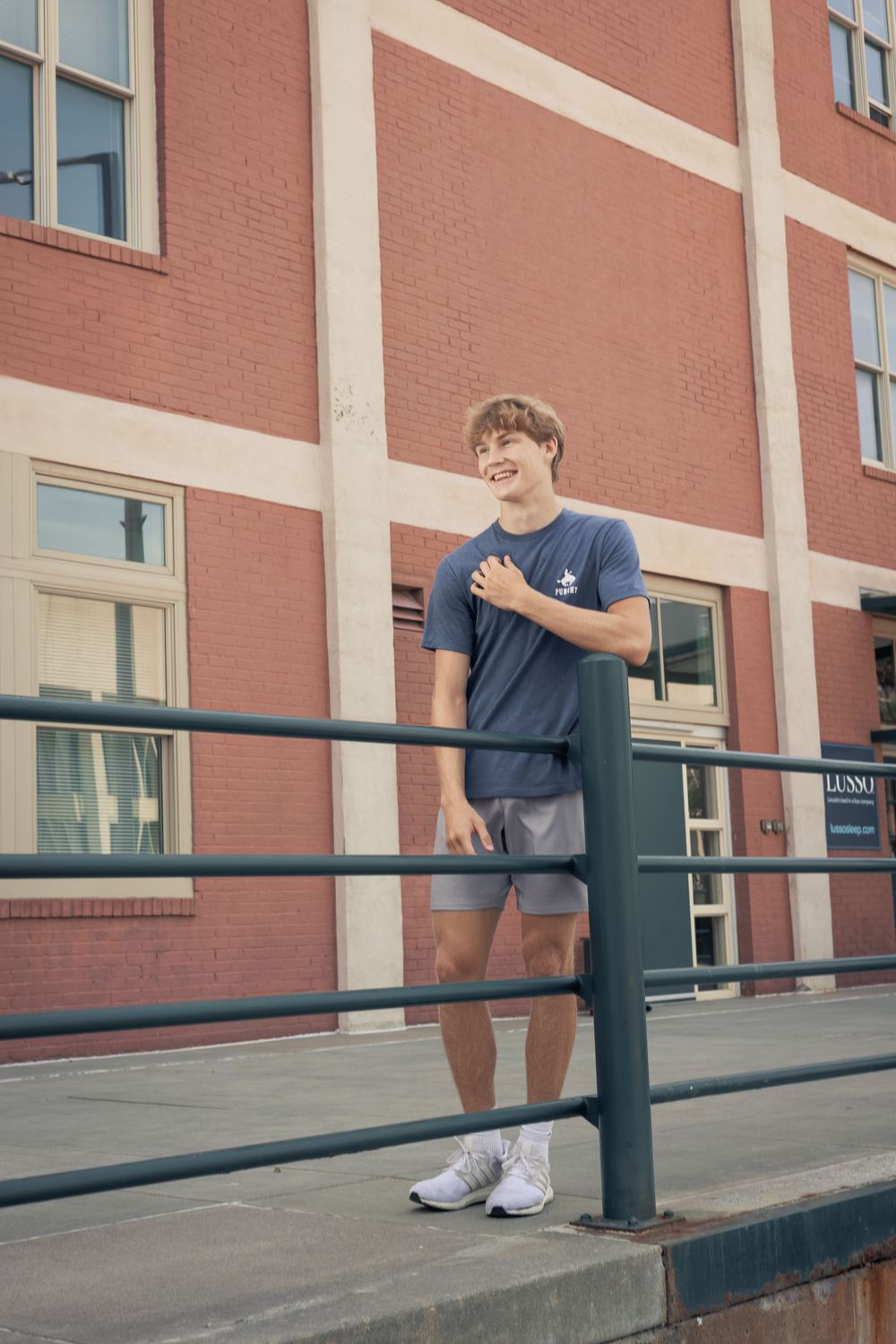 Person in casual attire leaning against a railing outside a brick building on a sunny day.