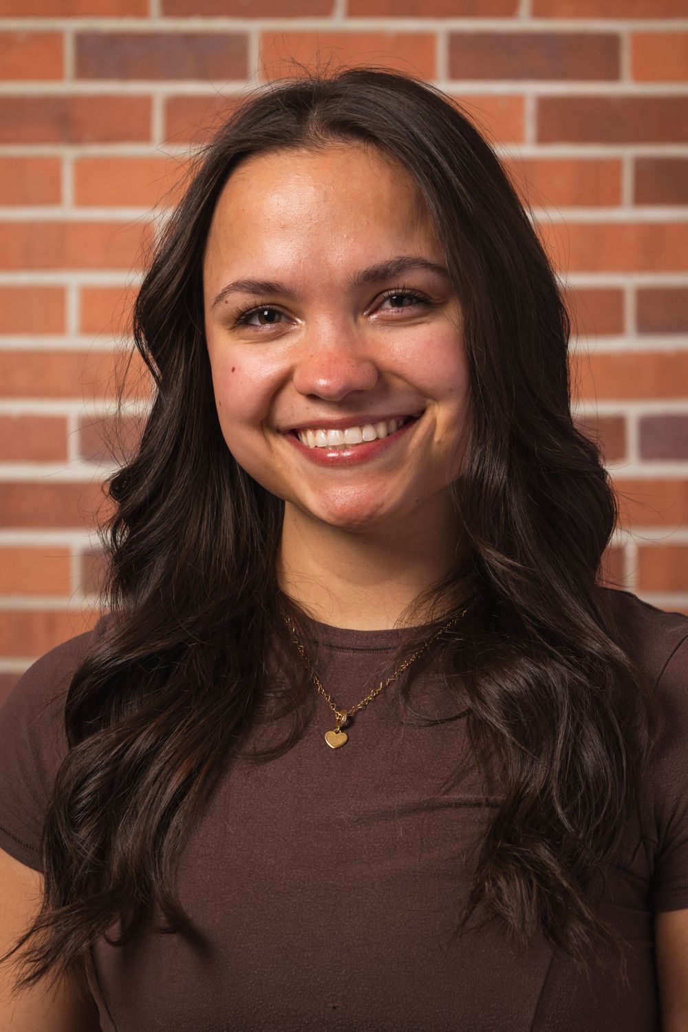 Portrait against a brick wall background wearing a brown top.