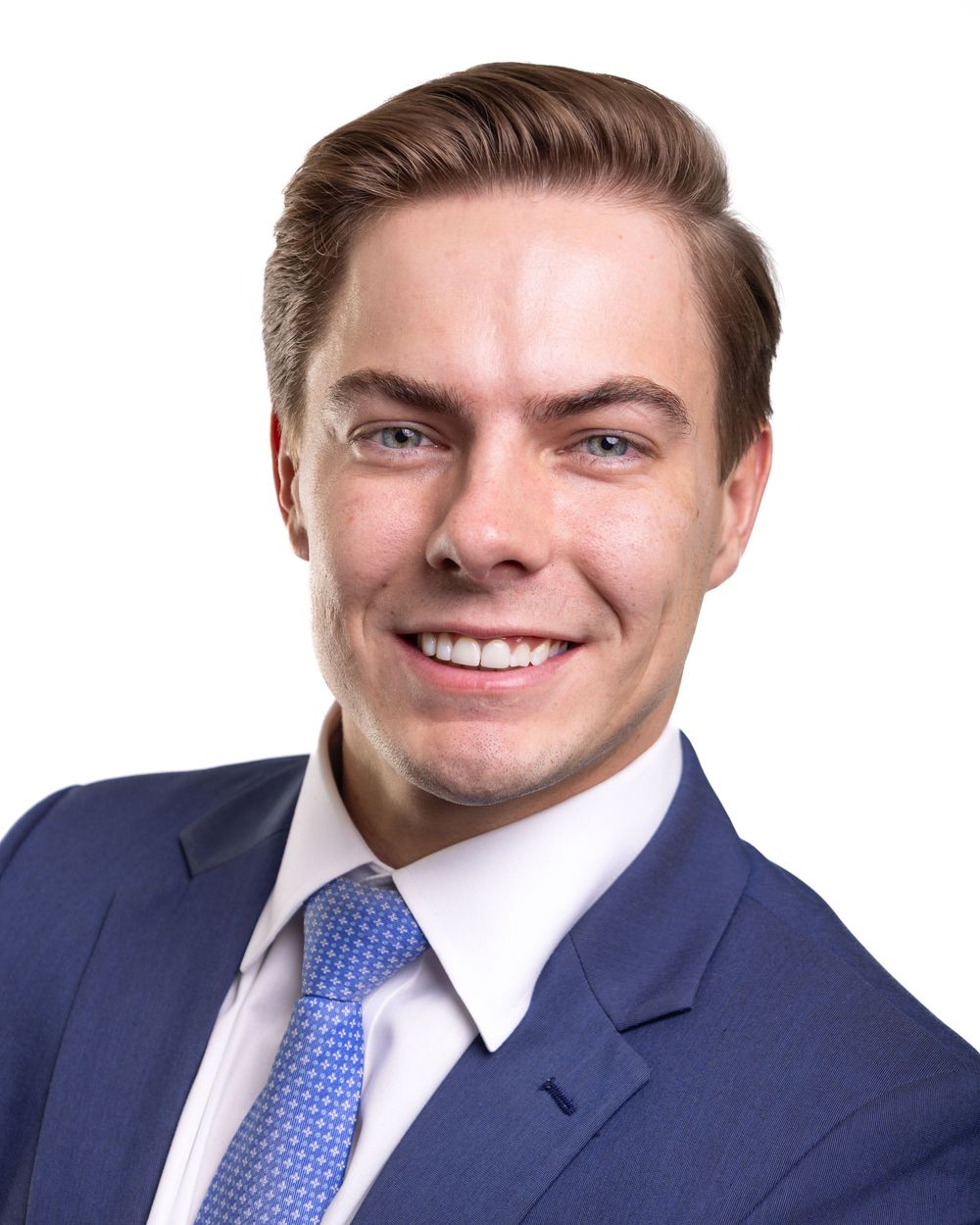 Professional headshot of a young businessman in a navy suit with blue patterned tie against white background.