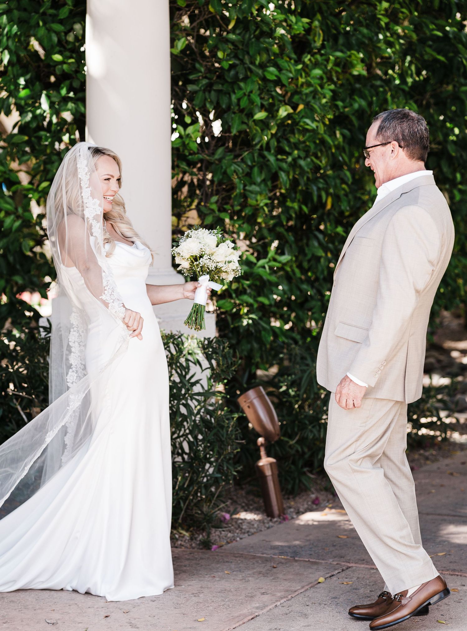 Bride and groom share a sweet moment under a white pillar surrounded by lush greenery at an outdoor wedding ceremony.