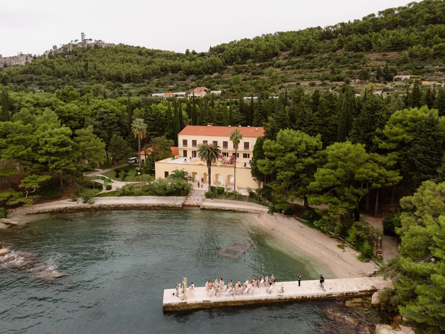 Aerial view of a pink Mediterranean villa with private beach and pier surrounded by pine trees.