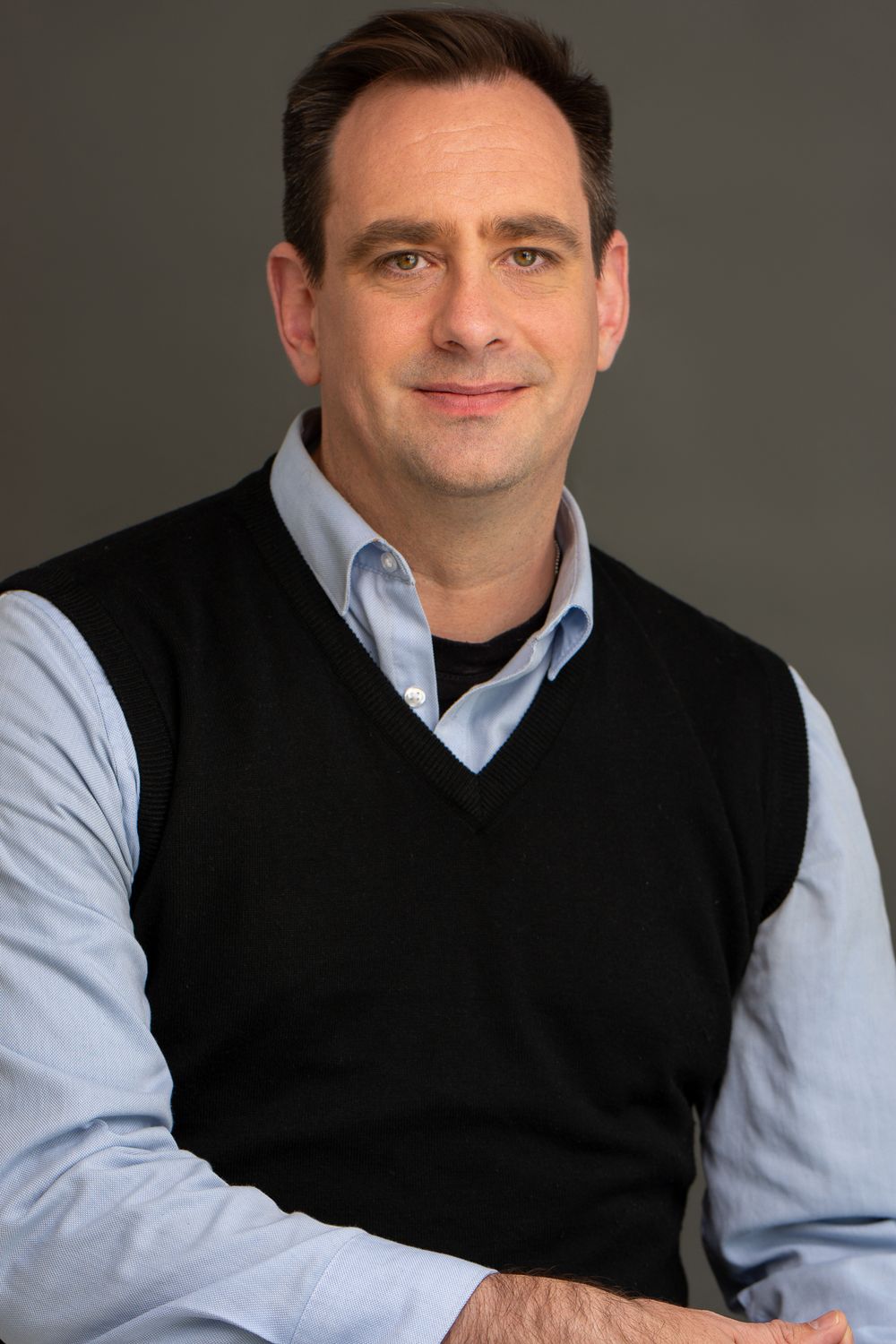 Corporate headshot of a man wearing a black sweater vest over a light blue button-down shirt in the studio setting in Cambridge, UK