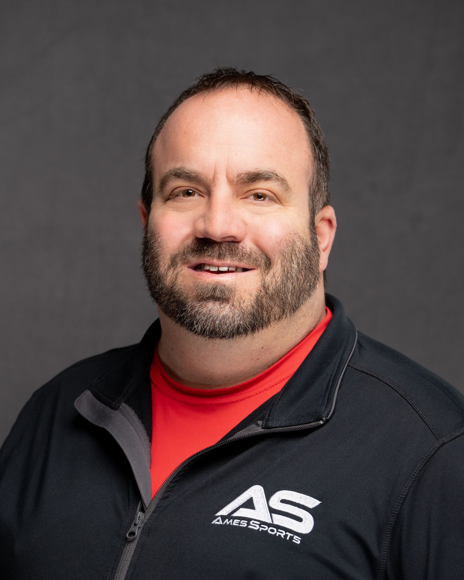 Professional headshot of person in red shirt and black jacket with AS logo against gray background.