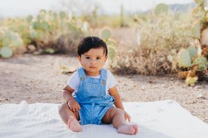Baby in blue overalls sits on white blanket during desert photoshoot.