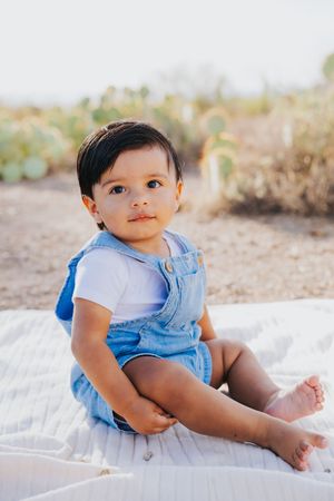 Infant wearing blue overalls sits on white blanket during outdoor desert photoshoot.