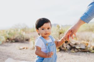 Baby in blue overalls reaches for parent's hand during desert photo session.