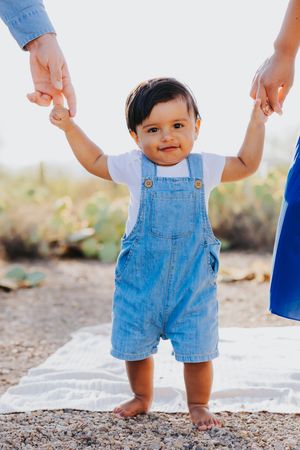 Baby in denim overalls stands supported by parents' hands during desert photoshoot.