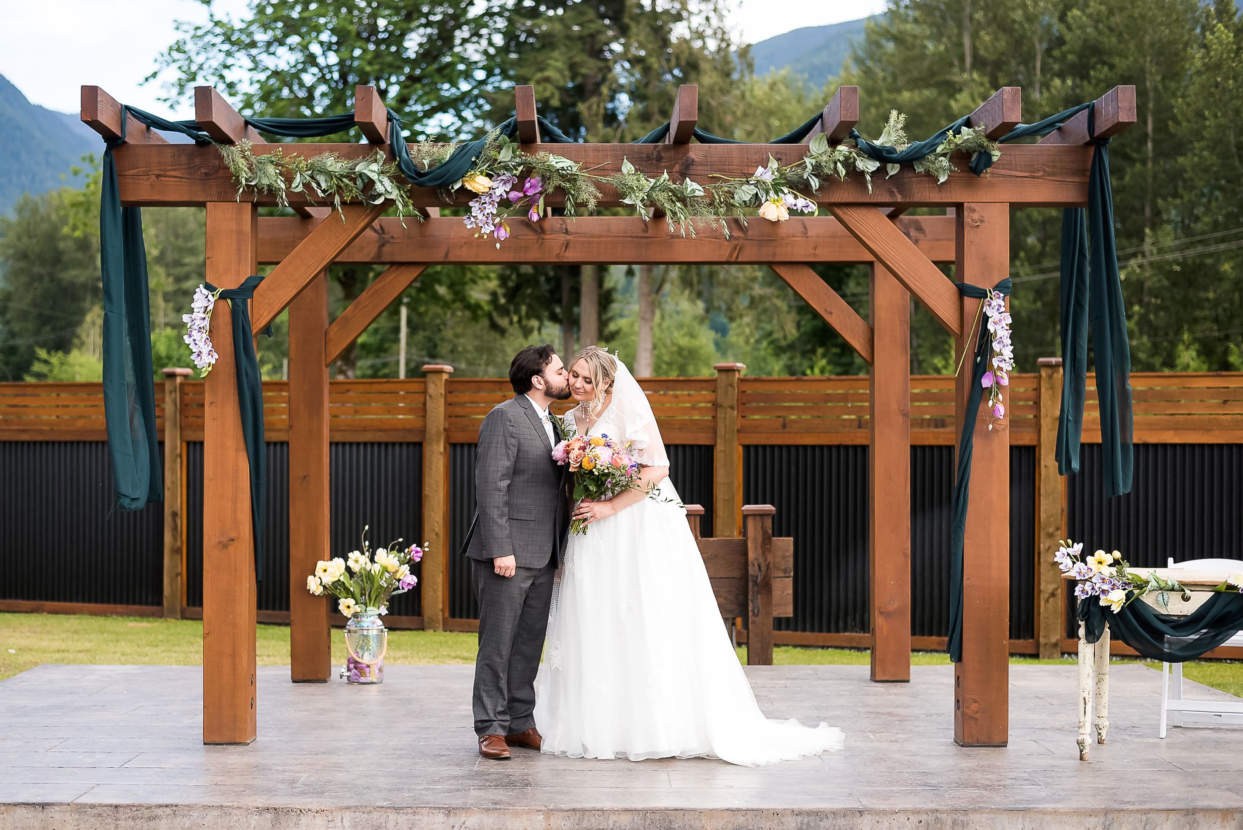 A groom kisses his bride on the cheek as they stand under their ceremony arch in Chilliwack, BC.