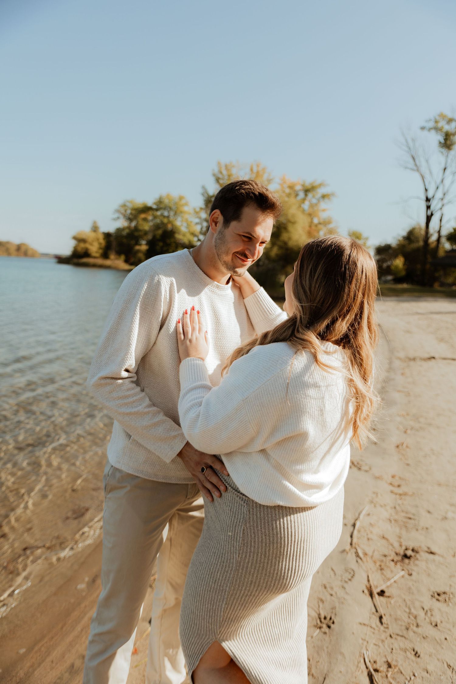 A couple in cream-colored sweaters embraces lovingly on a sunny beach with water and trees in the background.