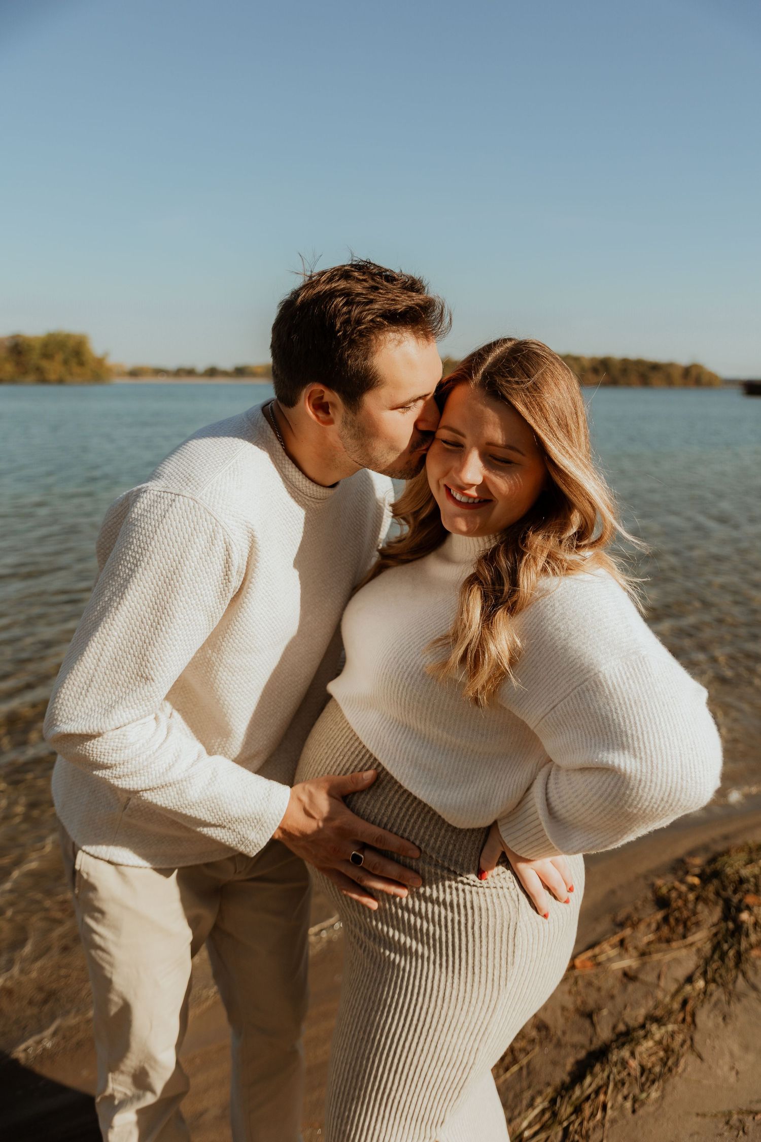 Couple in matching white sweaters embraces lovingly by a waterfront during golden hour.
