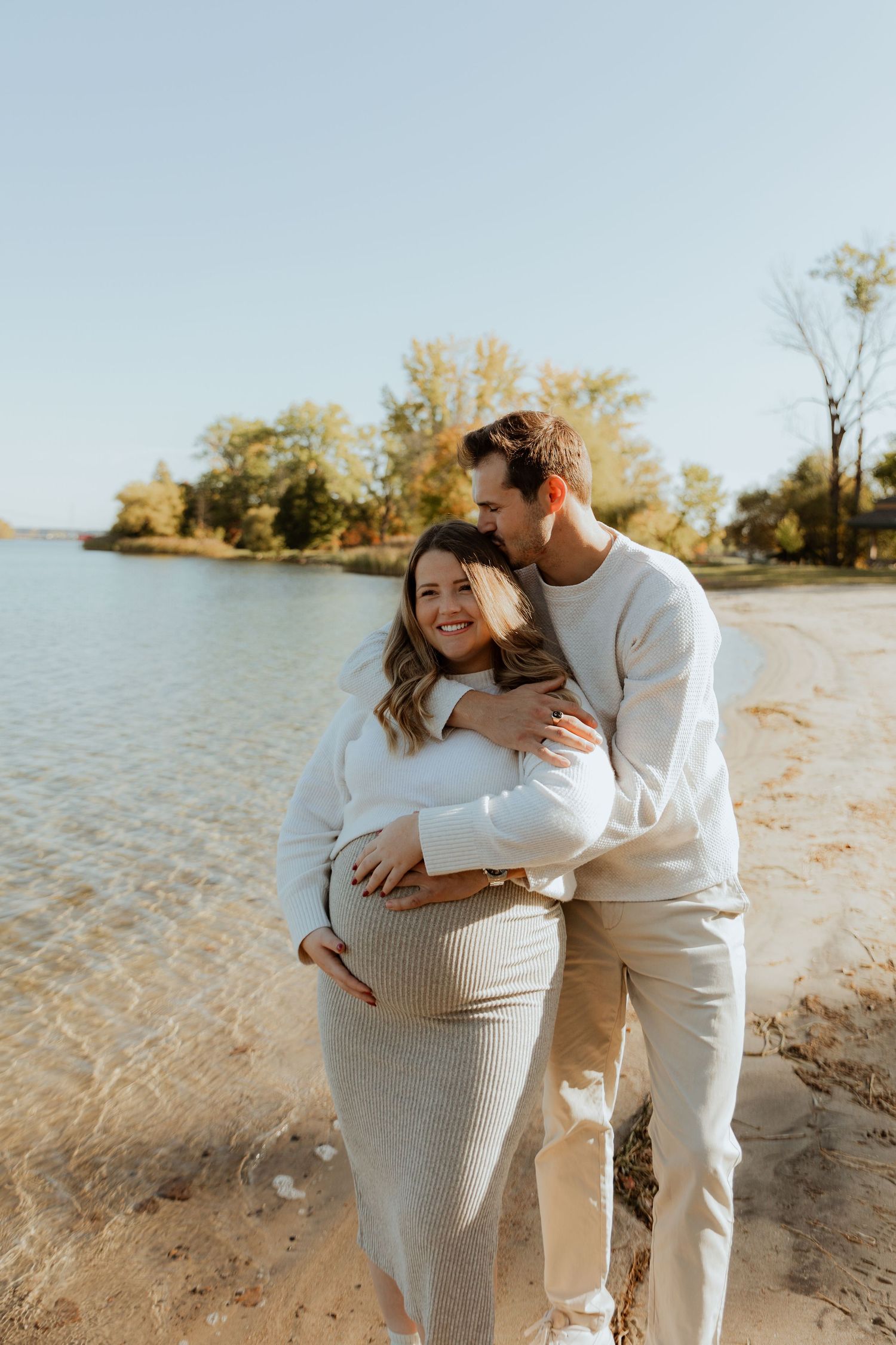 Couple embraces on a sandy beach during autumn with golden trees and water in the background.