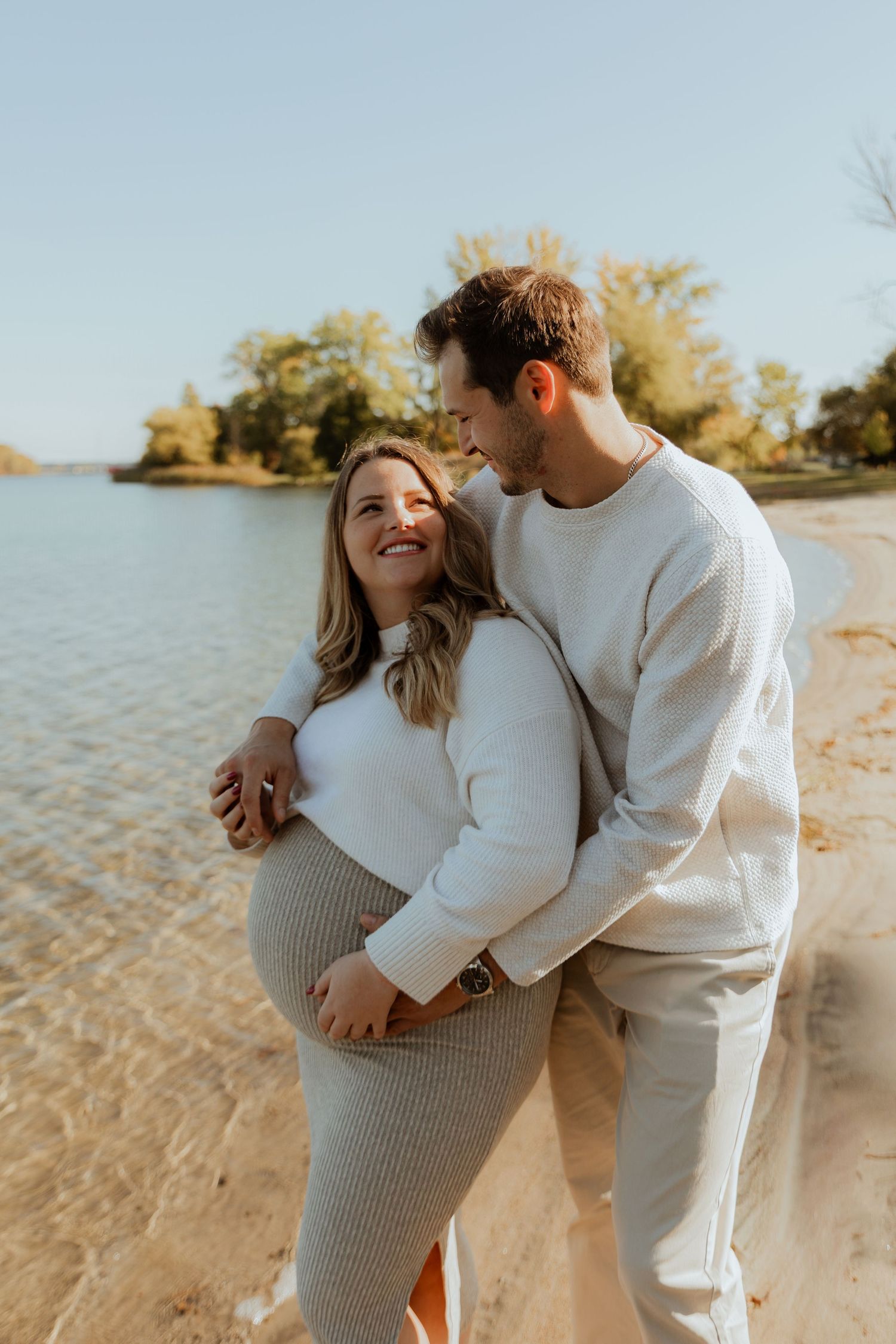 A couple in white and gray outfits share tender moments on a sandy beach during golden hour by the water.