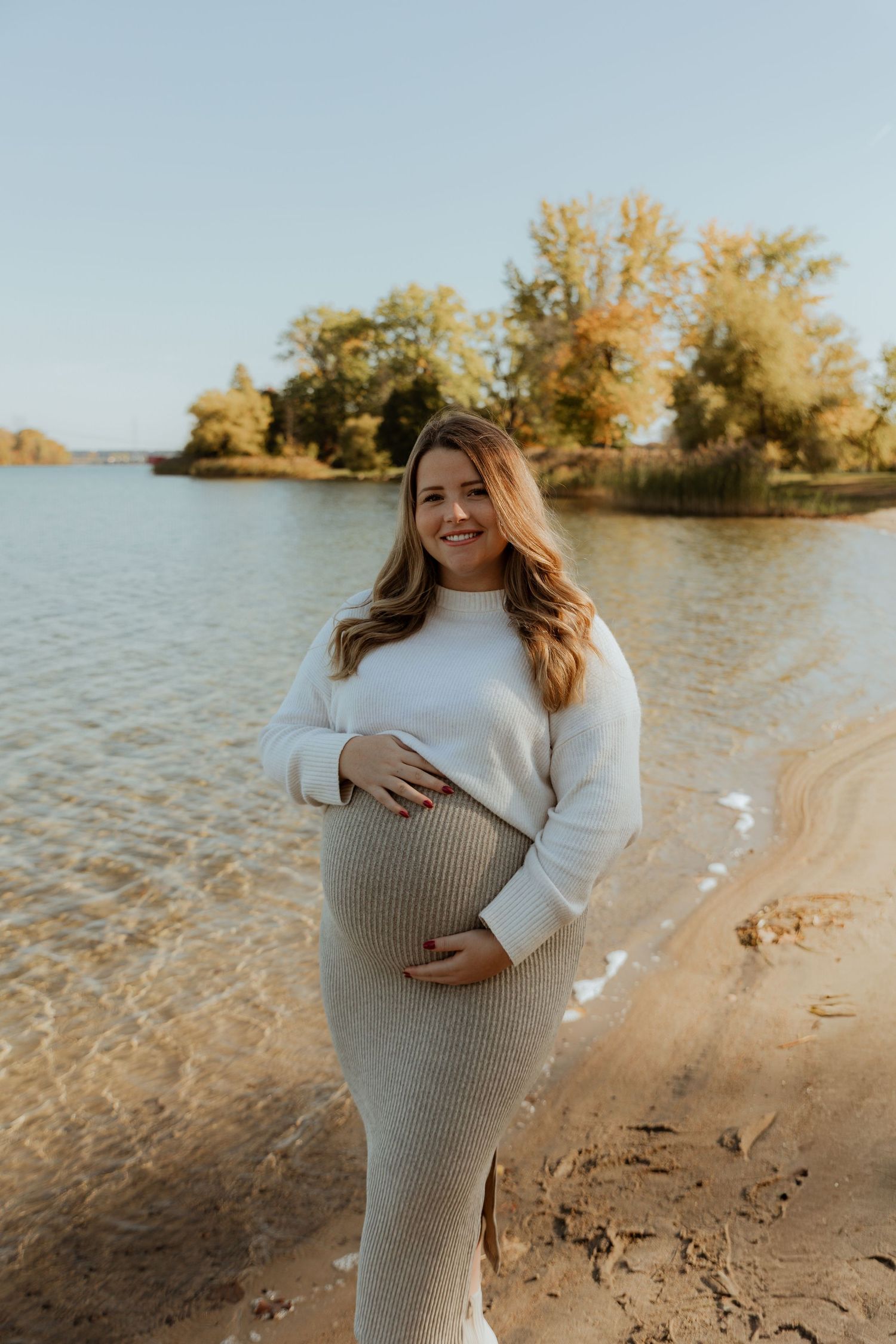 A maternity photoshoot at a beach during golden hour with fall foliage in the background.