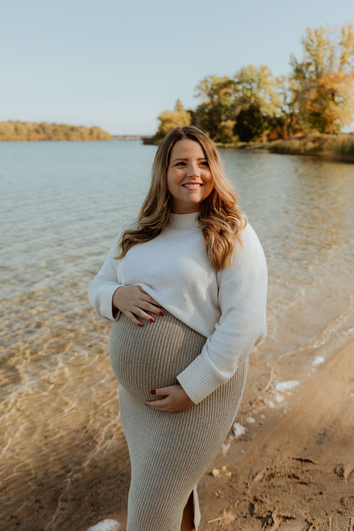 Maternity photoshoot at a lakeside beach during golden hour with autumn trees in the background.