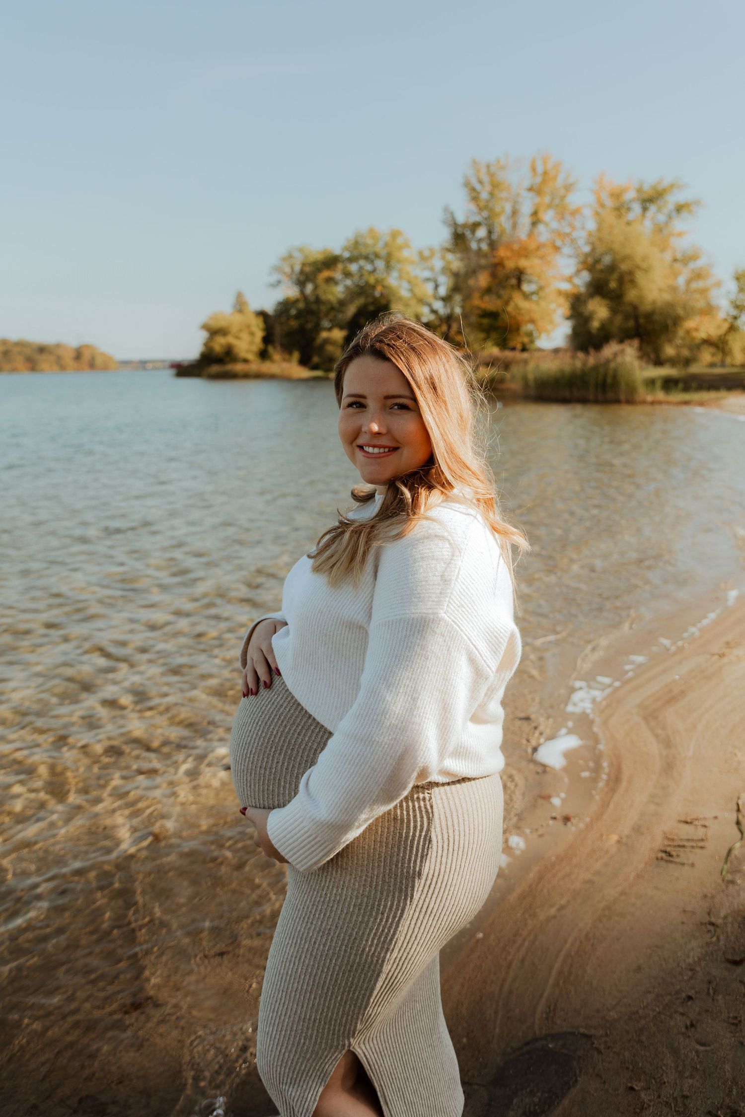 Expectant mother in white sweater poses on autumn lakeshore with golden trees in background.